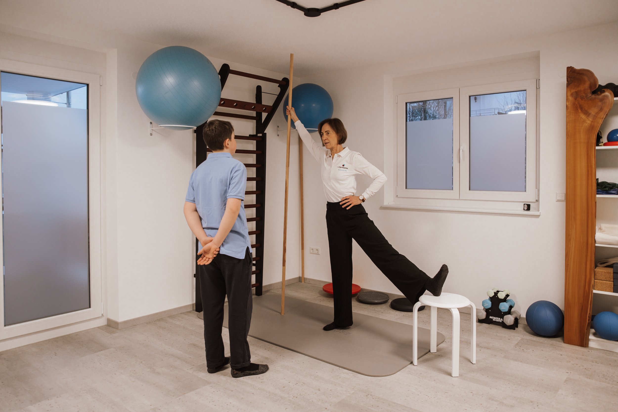 Frau beim Physiotherapie-Training mit einem jungen Patienten in einem Fitnessraum, der mit Gymnastikbällen und anderem Trainingszubehör ausgestattet ist.