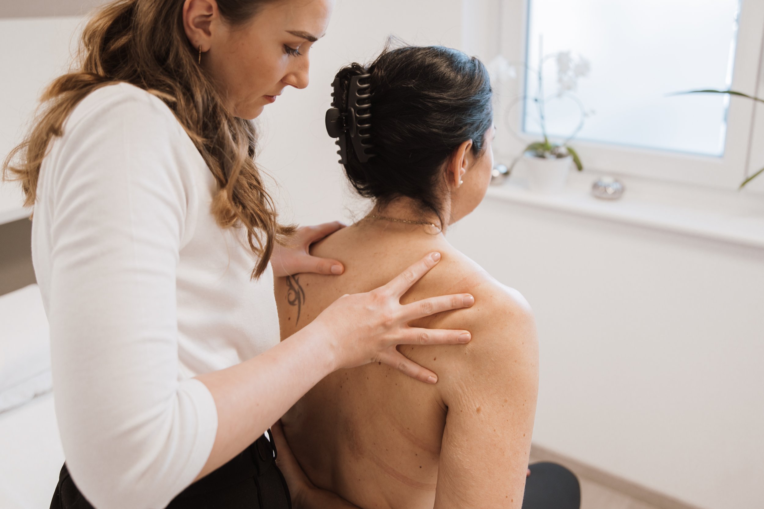 Ein Therapeut behandelt eine Frau im Stehen, die auf einem Tisch sitzt, mit den Händen auf der Schulter und dem Rücken. Die Frau trägt eine schwarze Haarspange.
