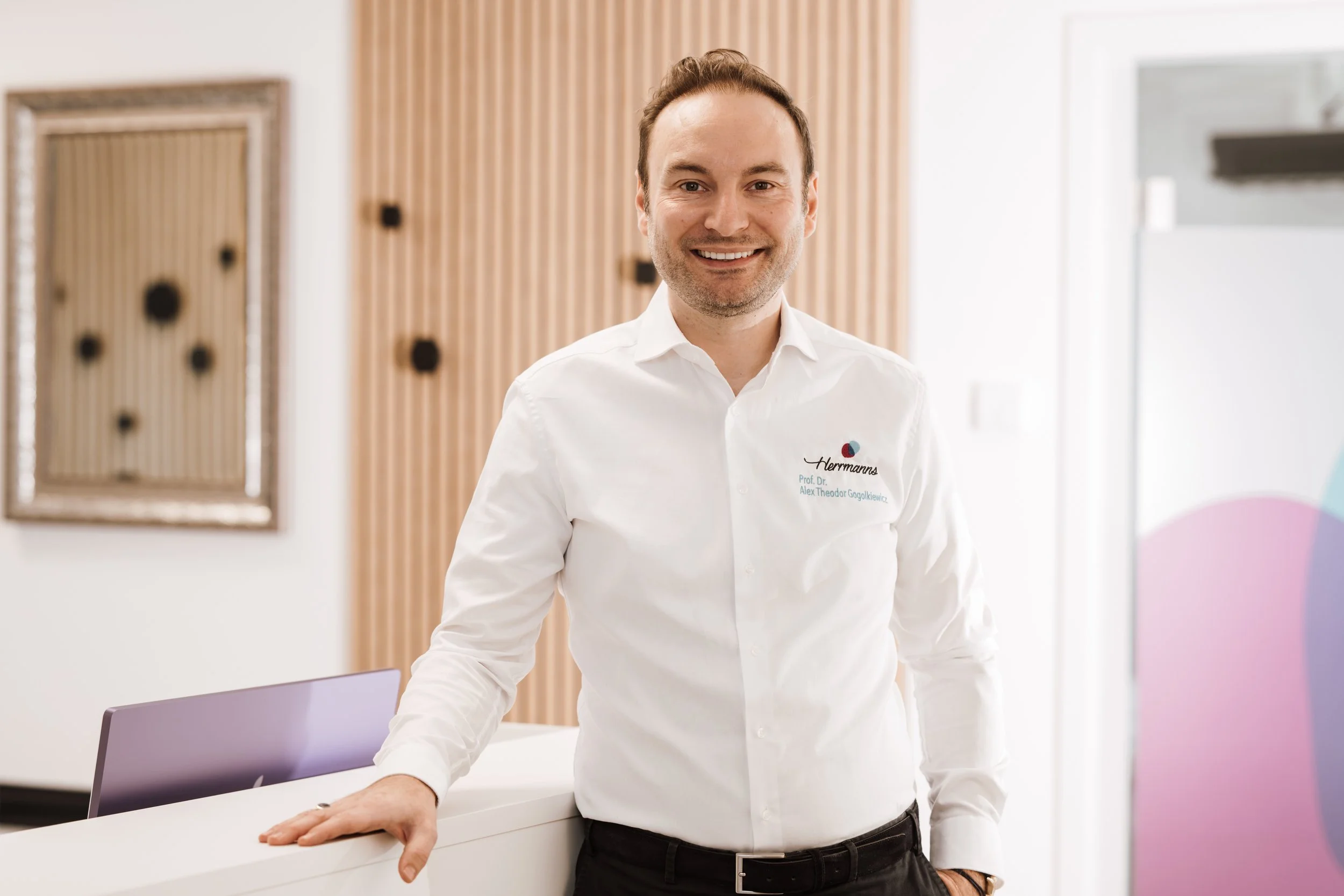Ein lachender Mann in weißen Hemd, der an einer Rezeption lehnt, in einem modernen, hellen Raum. Das Hemd trägt ein Logo sowie den Namen Prof. Dr. Alex Theodor Gogalkiewicz.