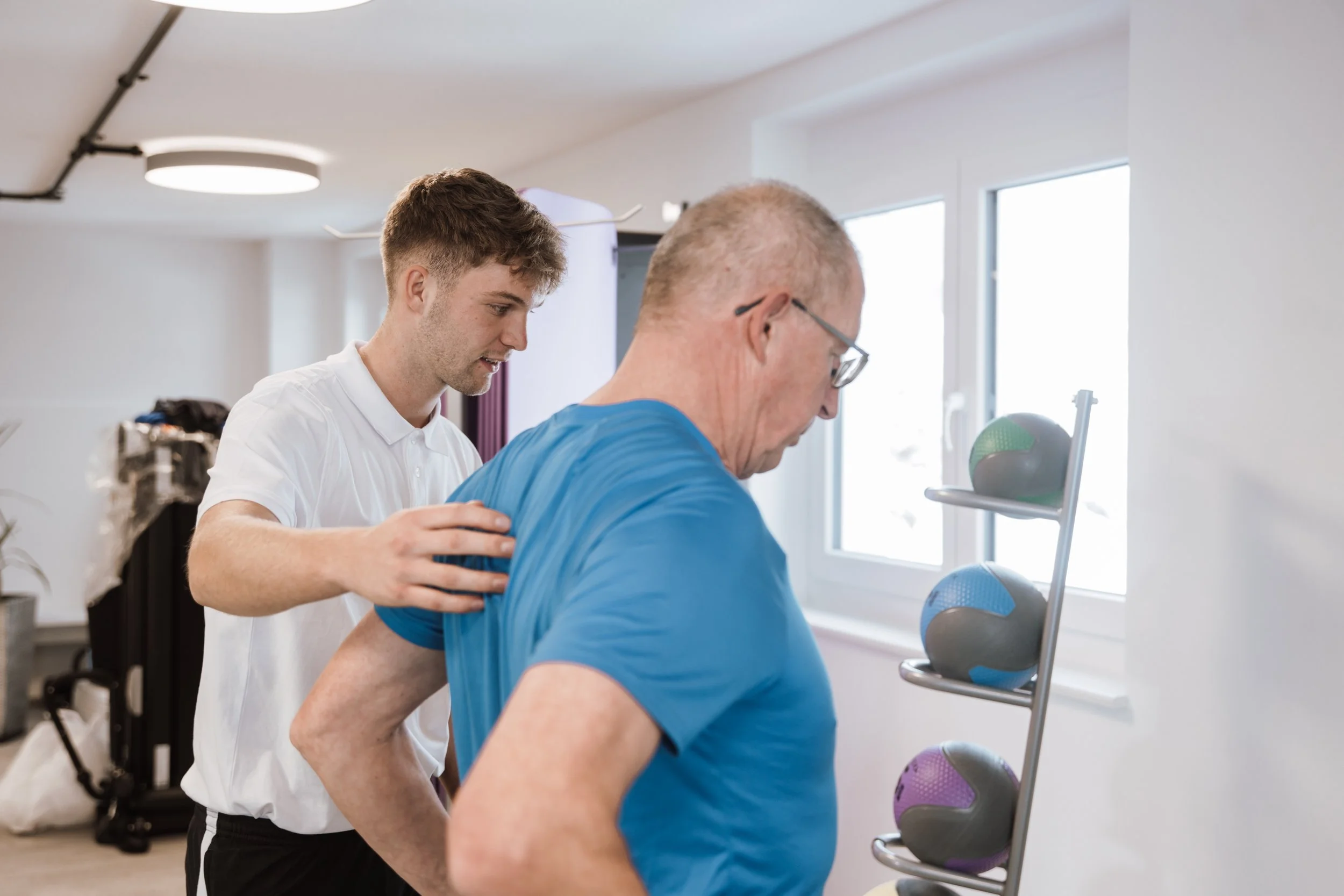 Ein junger Mann hilft einem älteren Mann, seine Schulter zu behandeln, in einem Fitnessraum mit Medizinbällen auf einem Regal neben dem Fenster.