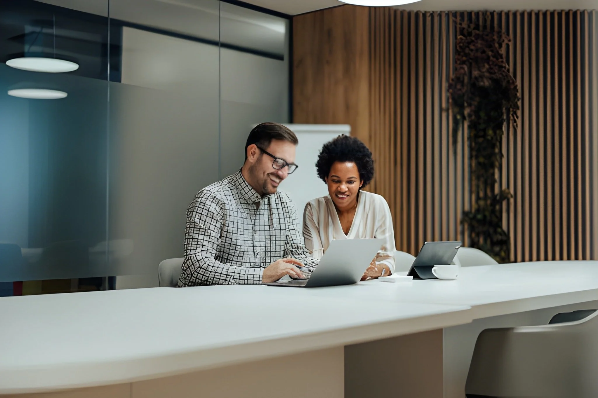Two diverse professionals, a man and a woman, are sitting at a conference table working on a laptop and a tablet in a modern office space.