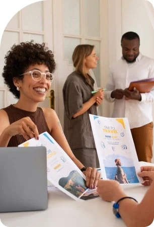 Three people at a table, two women and one man, discussing travel brochures; the woman in the foreground is smiling and holding travel flyers, with a laptop nearby.
