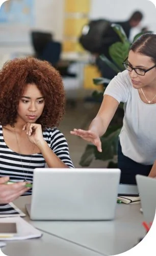 A woman appears to be scolding or explaining to a young girl sitting at a desk with a laptop, in an office setting.