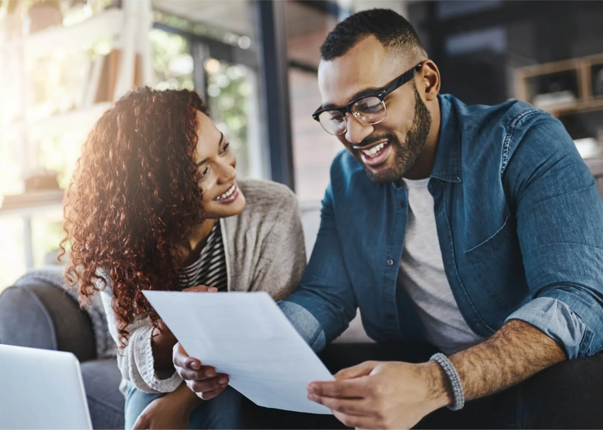 A man and woman sitting close together, looking at a piece of paper and smiling, in a cozy, well-lit room.