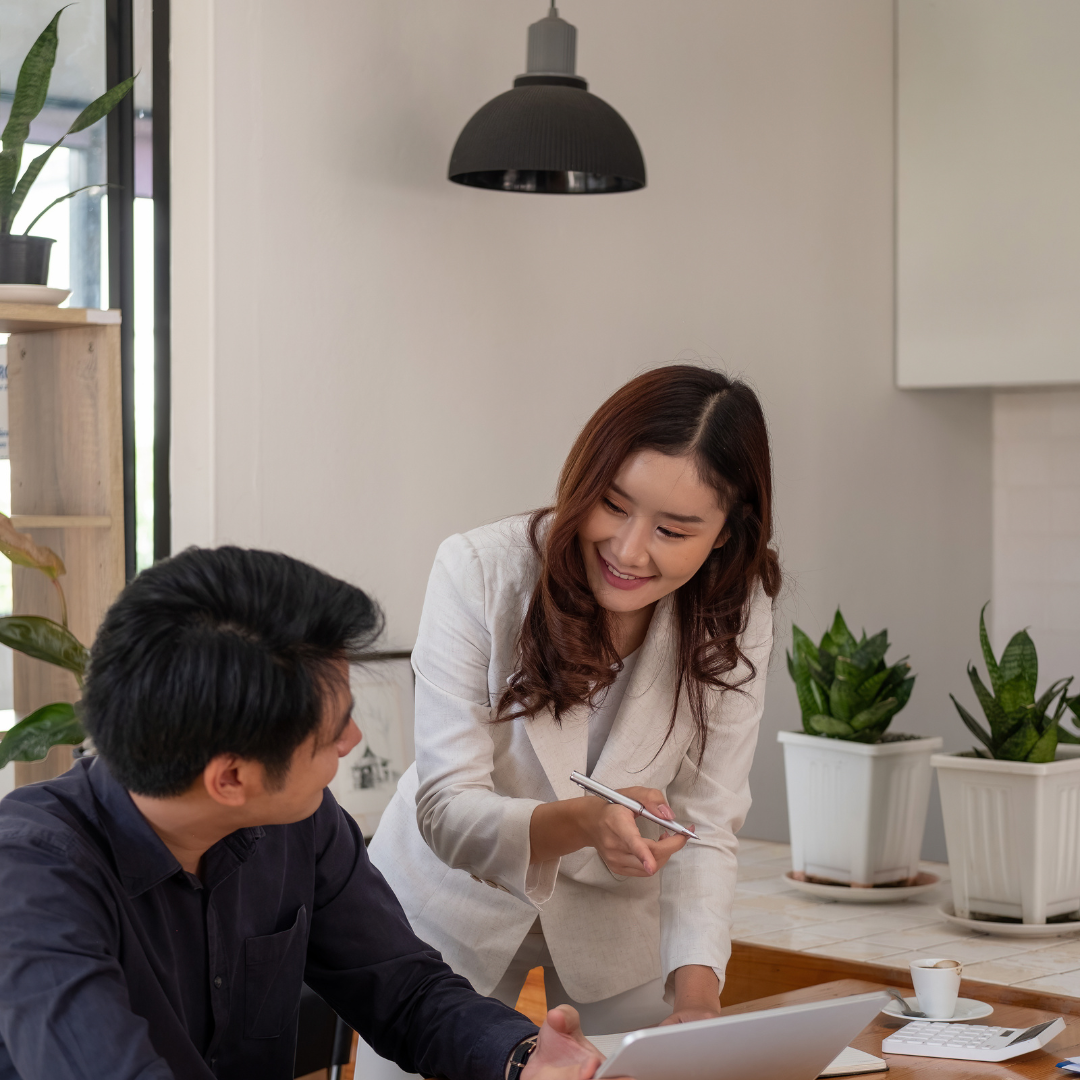 A woman in a cream blazer is smiling and leaning over a desk, talking to a man in a dark shirt who is sitting and looking at a laptop. The setting is a modern, well-lit room with plants and decorative elements.