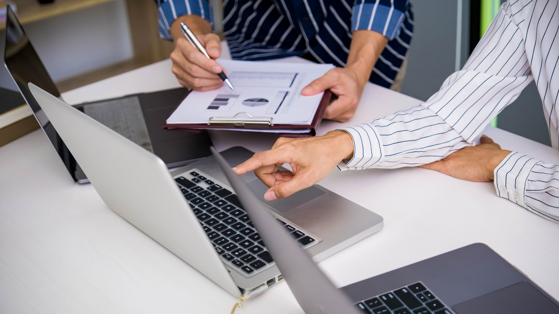Two individuals sit at a table with laptops, one pointing at the laptop screen and the other holding a clipboard with documents and graphs.