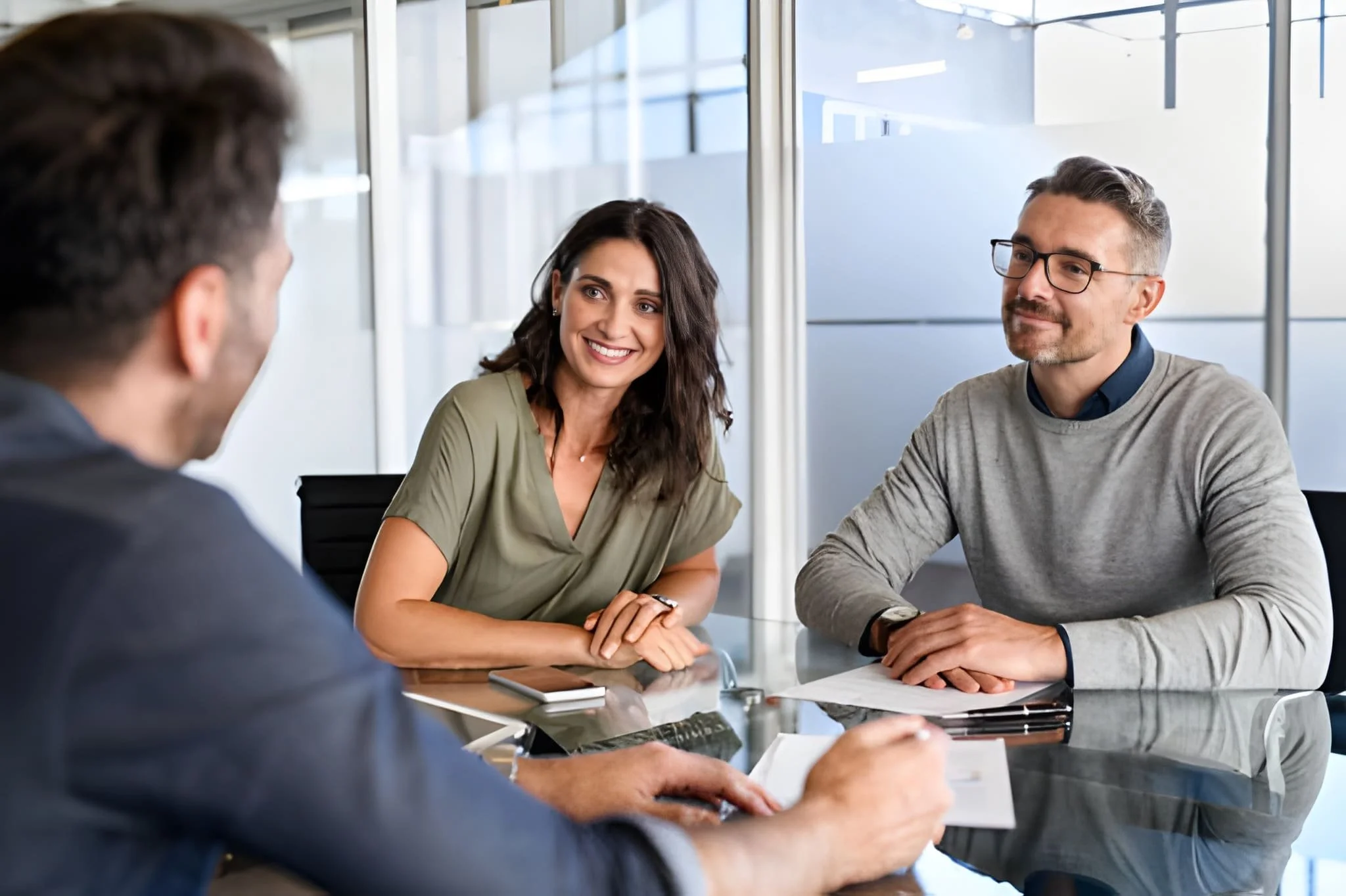 Three people sitting at a glass conference table engaged in a discussion, with a woman smiling and two men listening in a modern office setting.