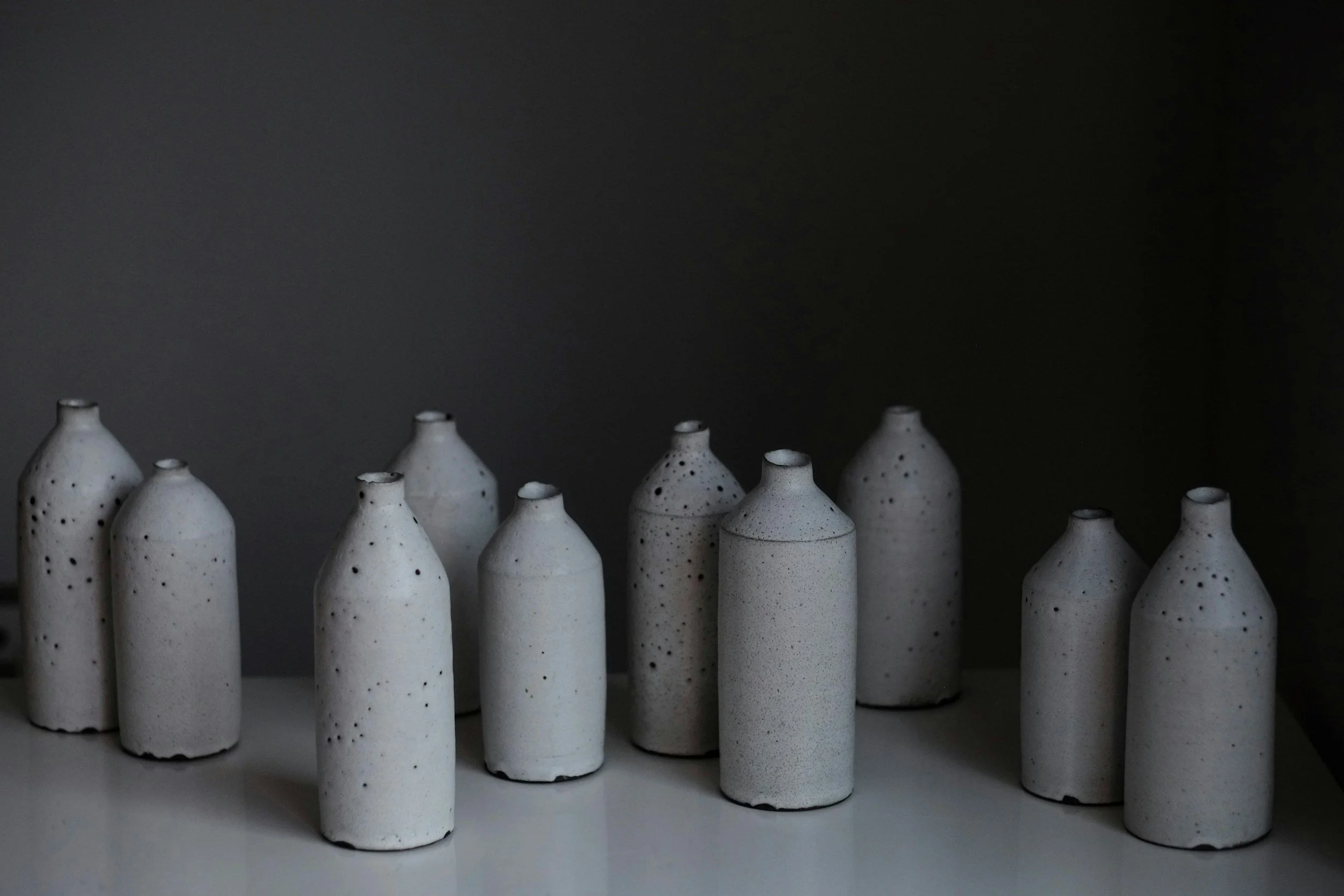 A collection of ten white ceramic vases with black speckles arranged in a row on a white surface against a dark background.
