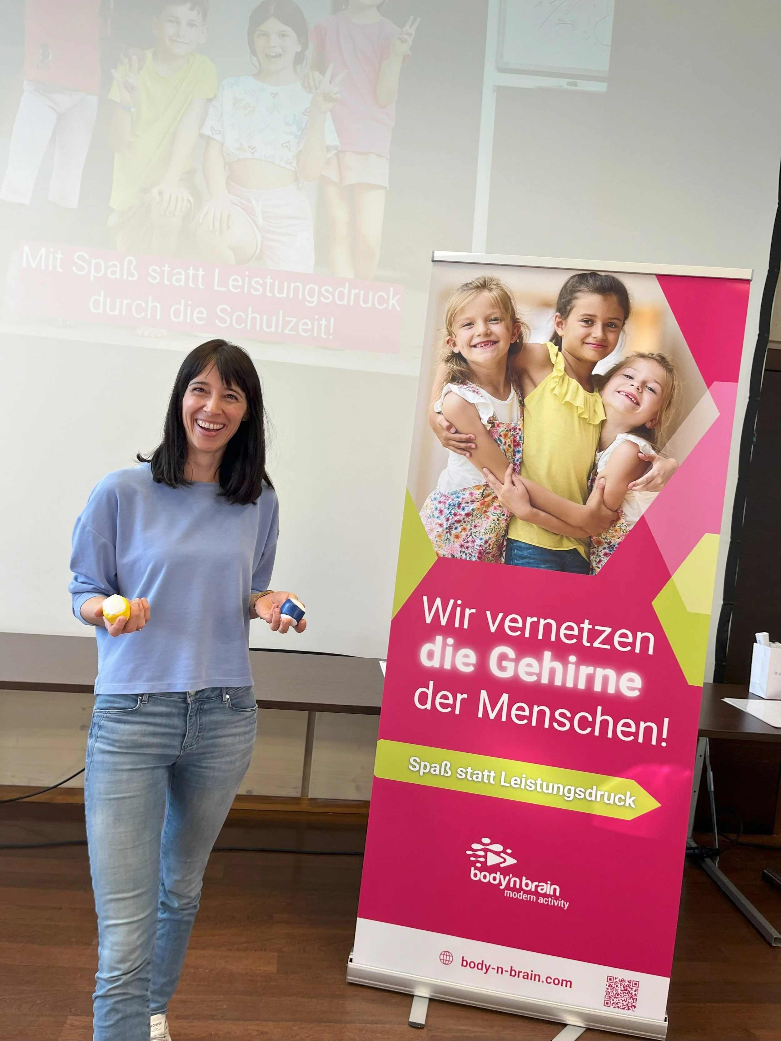 Eine Frau steht neben einem bunten Plakat, auf dem Kinder abgebildet sind. Das Plakat trägt den Text "Wir vernetzen die Gehirne der Menschen! Spaß statt Leistungsdruck" und zeigt eine Gruppe von lachenden Kindern.
