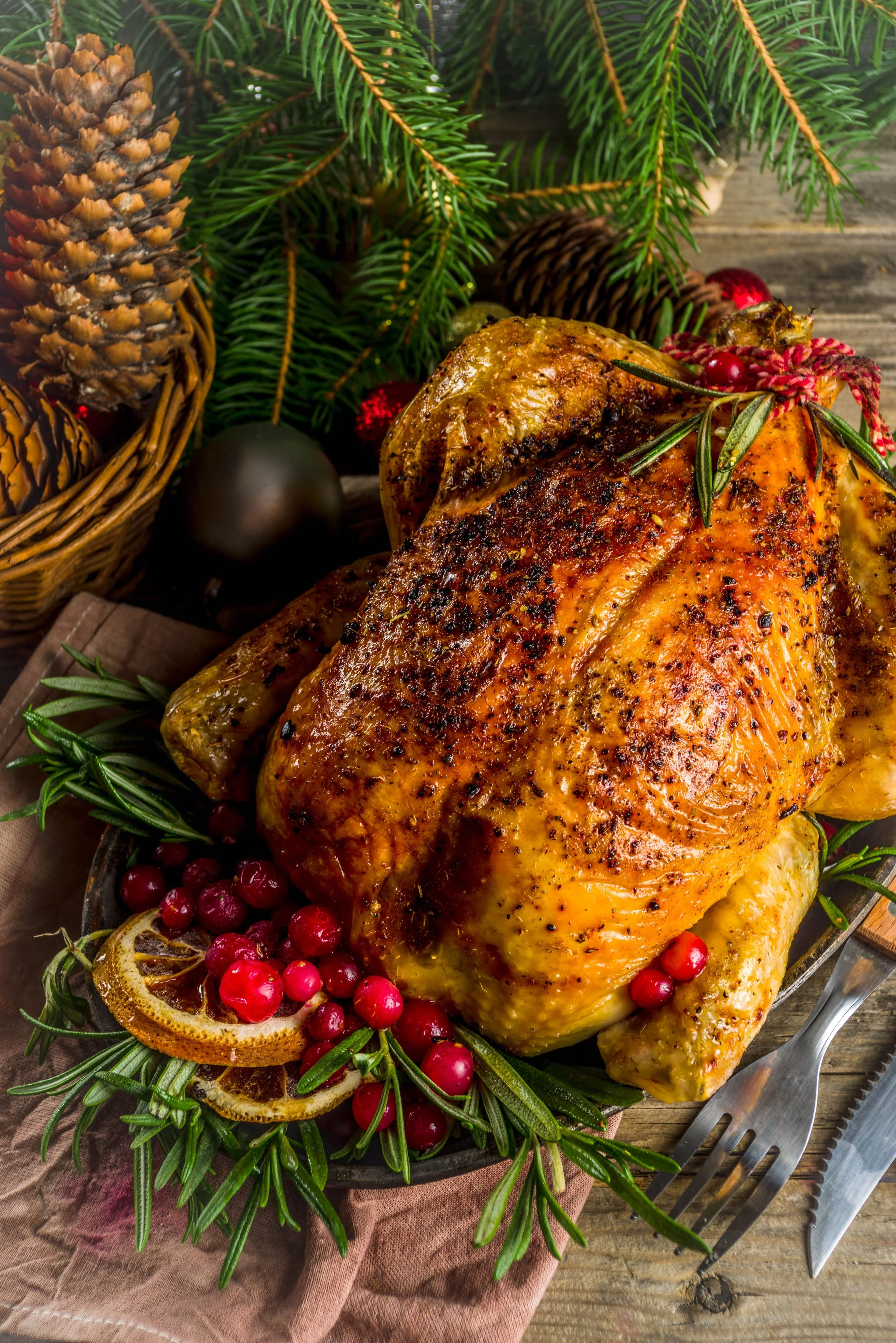 A roasted whole turkey garnished with lemon slices, cranberries, and fresh herbs, surrounded by pinecones, evergreen branches, and Christmas ornaments on a wooden table.