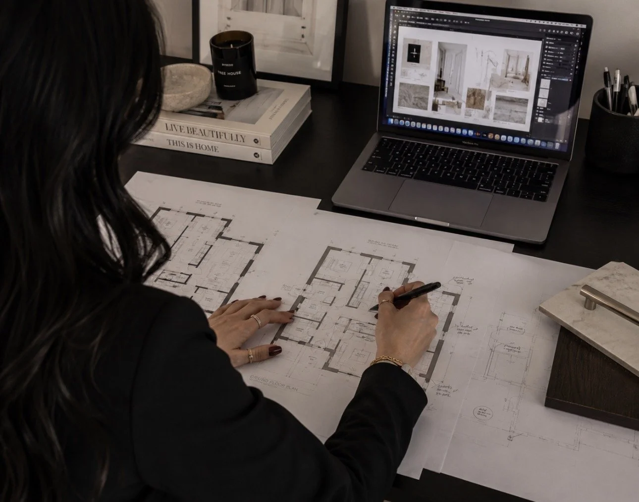 A woman working on architectural blueprints at her desk, with a laptop displaying interior designs, surrounded by books and office supplies.