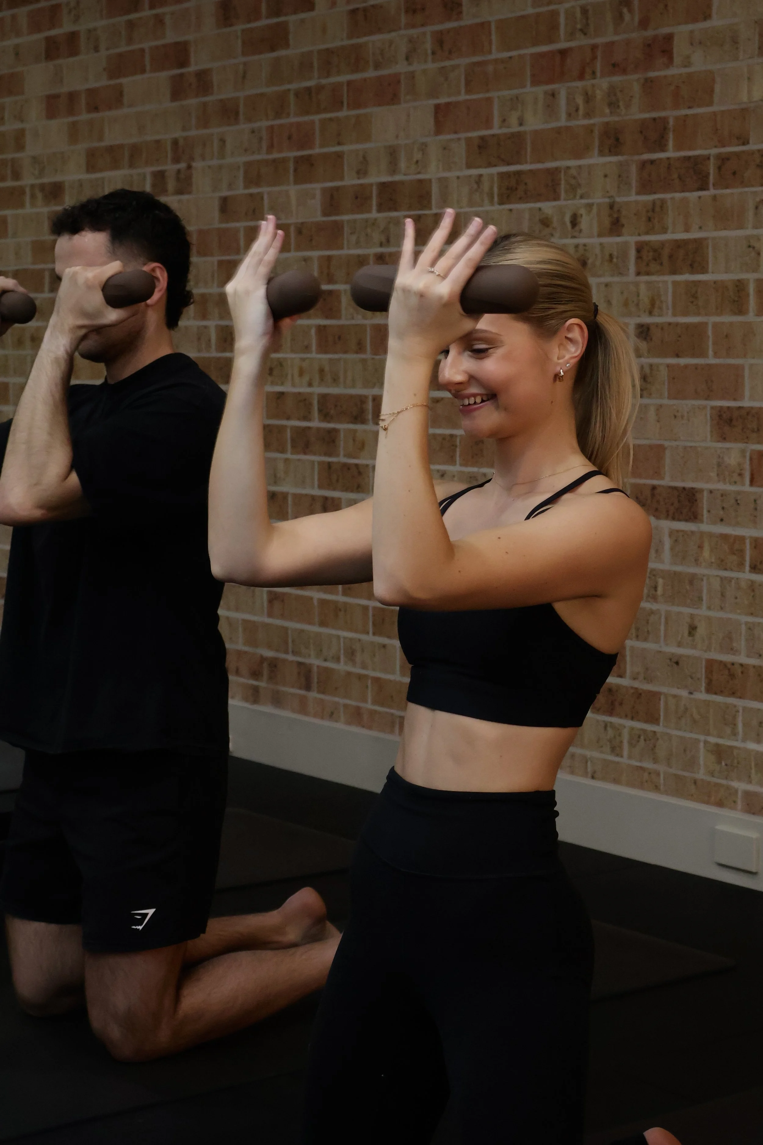 Two people are practicing pilates, kneeling on mats against a brick wall, holding foam roller bars above their heads with their hands,  and smiling.