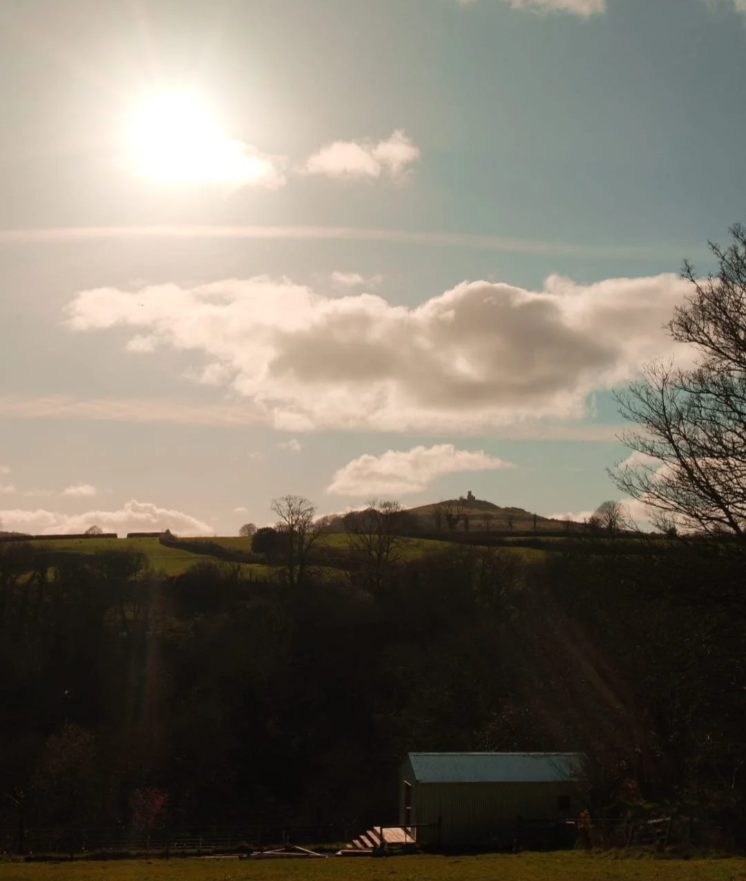 Landscape of rolling hills with trees and a small building in the foreground, under a partly cloudy sky with the sun shining brightly.