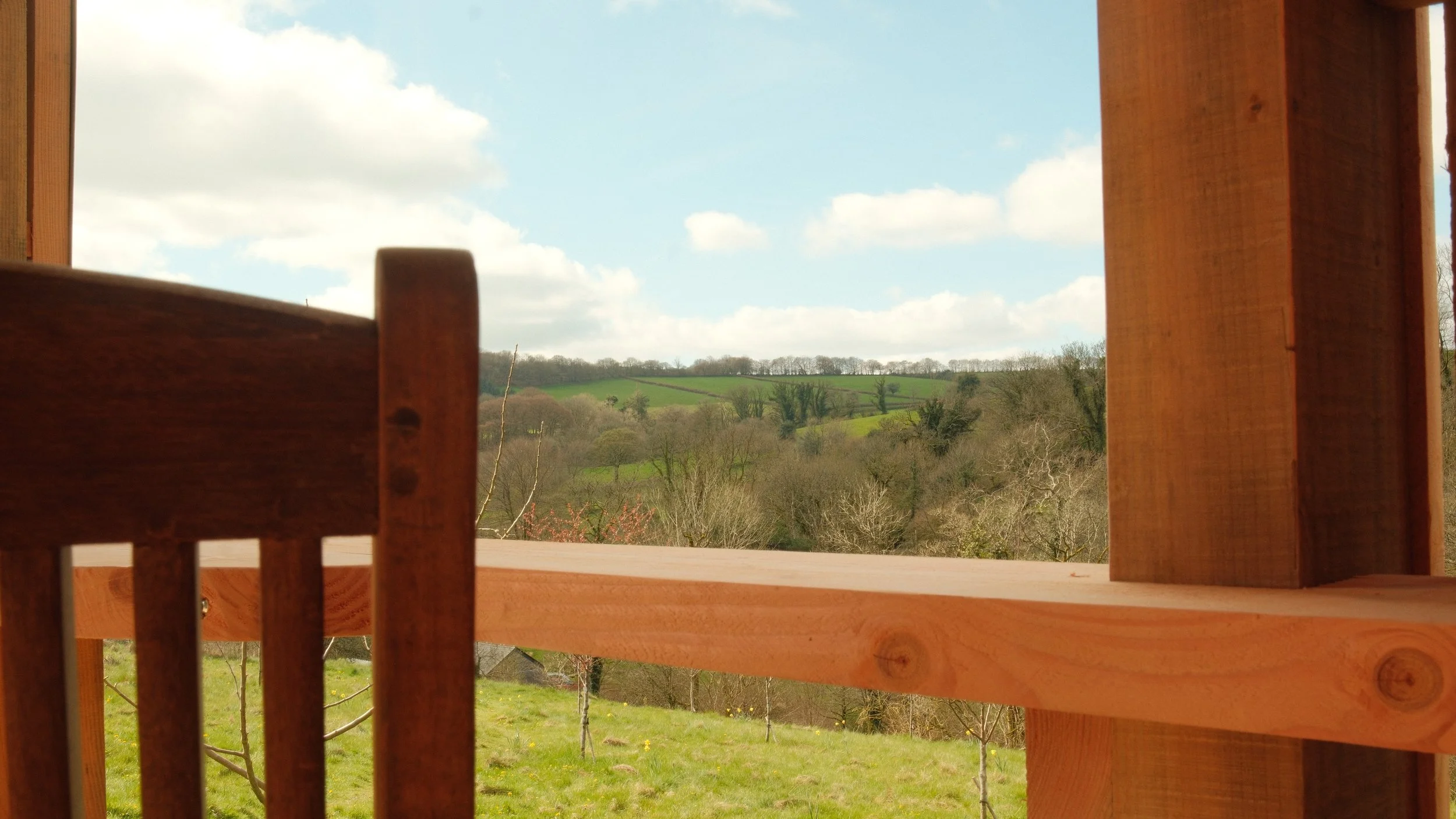 View through a wooden window frame, showing a rural landscape of rolling green hills with trees and a partly cloudy sky.