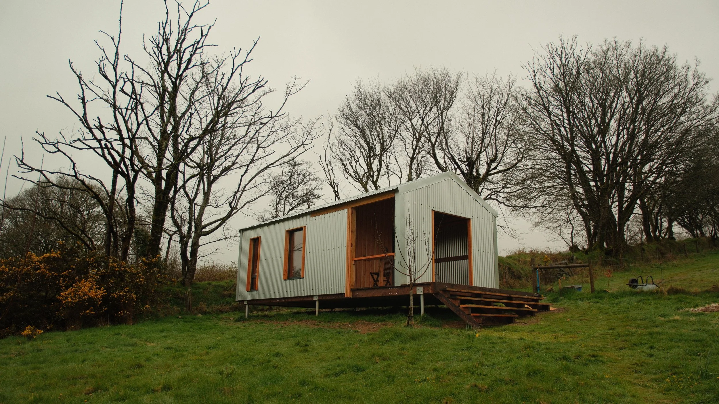 A small, metal shed on stilts with a wooden deck and stairs, surrounded by green grass, trees, and bushes on a cloudy day.