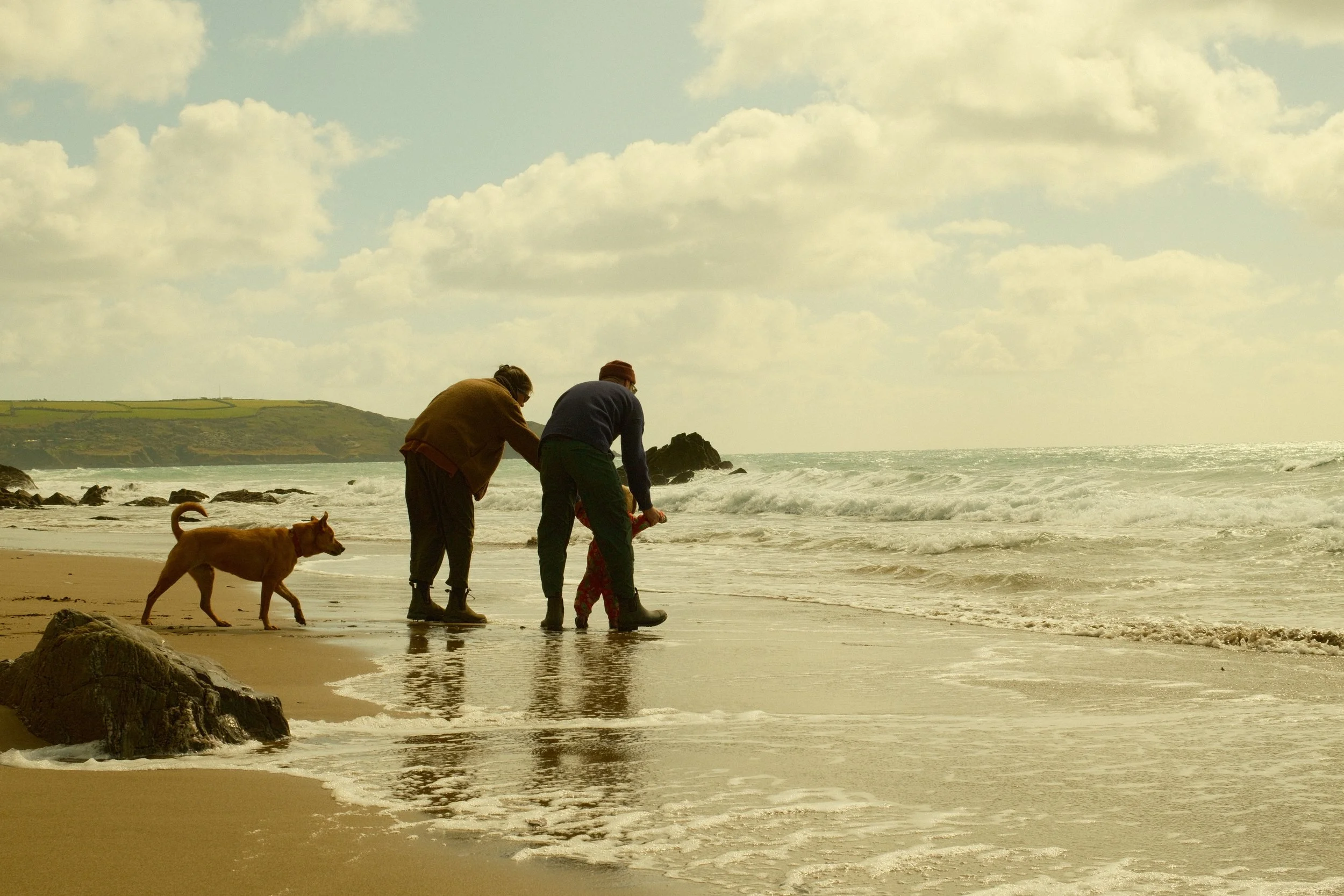 Two people and a dog on the beach, with waves and cliffs in the background under a partly cloudy sky.
