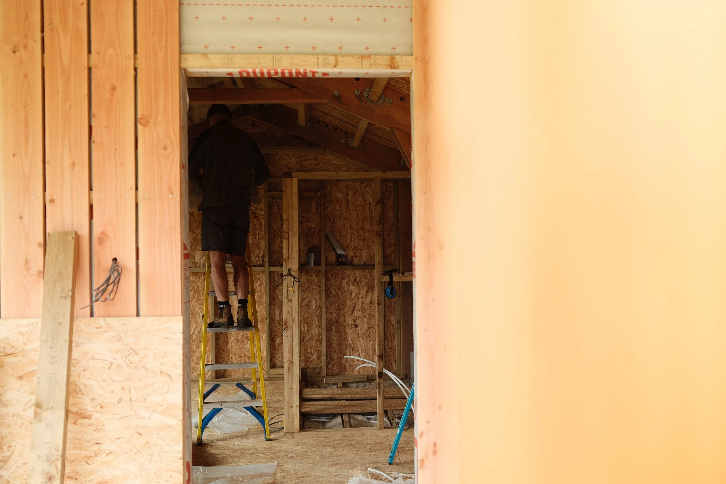 A worker standing on a yellow ladder inside a wooden framed room under construction, with wooden walls and ceiling, and construction materials around.