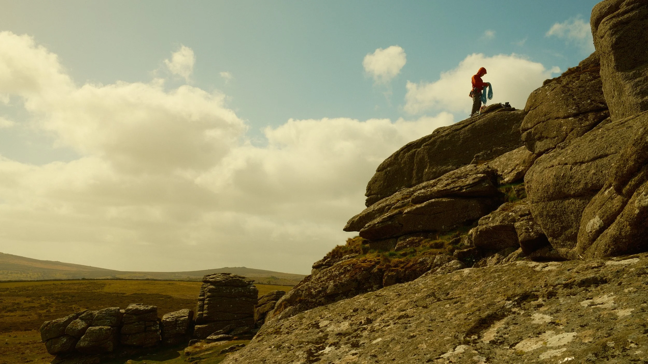 A person standing on a rocky cliff holding climbing gear, overlooking a vast landscape with grassy plains and distant hills, under a partly cloudy sky.