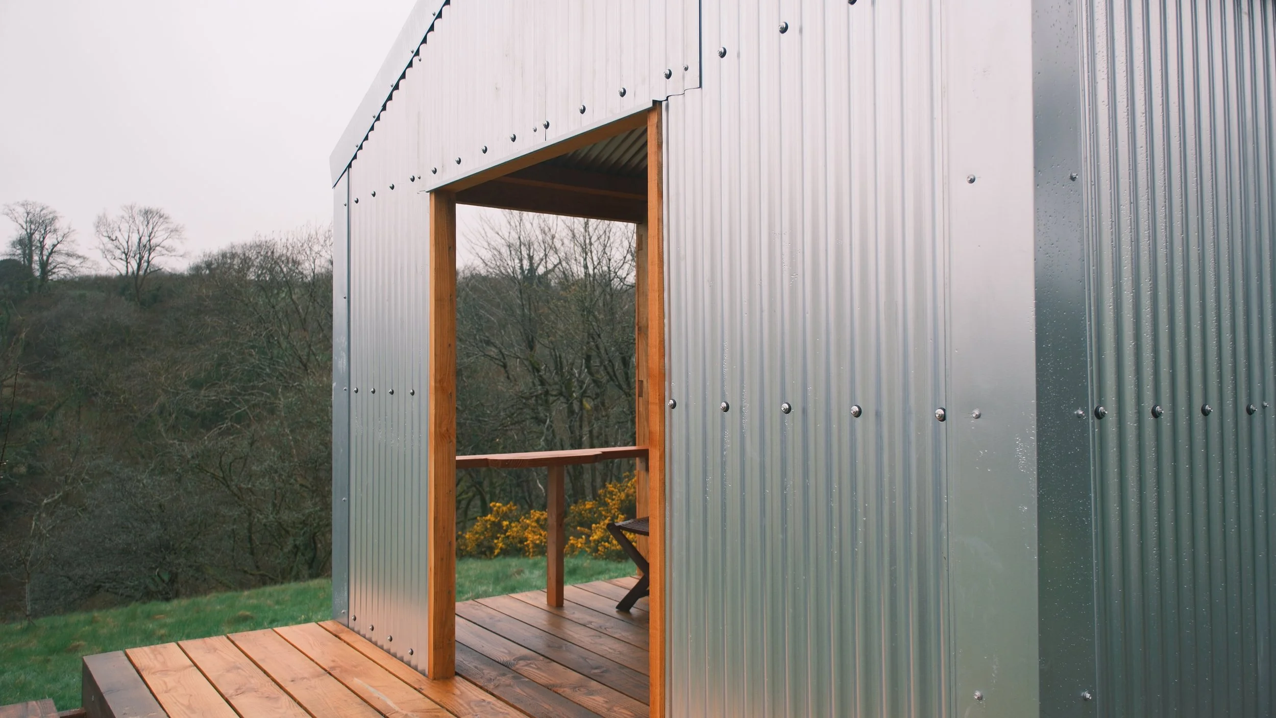 Steel building under construction with open side revealing wooden frame and balcony, set in a rural area with trees and grass