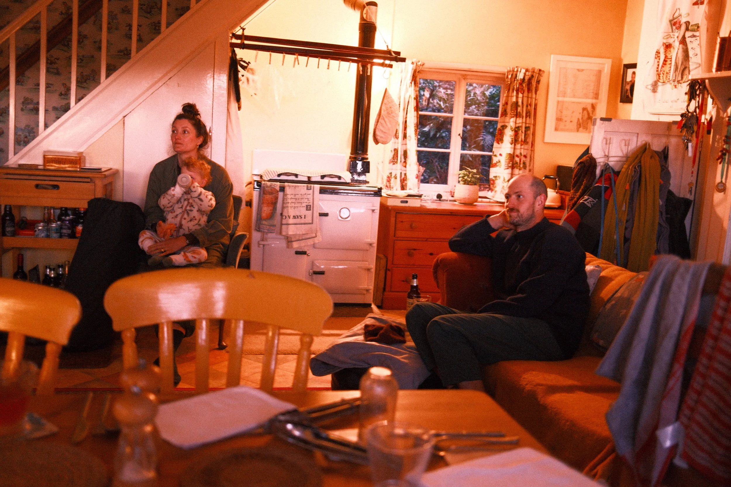 A cozy living room with two adults and a young child sitting and relaxing. The woman has her hair in a bun, holding the child who is drinking from a cup. The man has a beard, sitting on a chair, resting his head on his hand. The room has a window wit