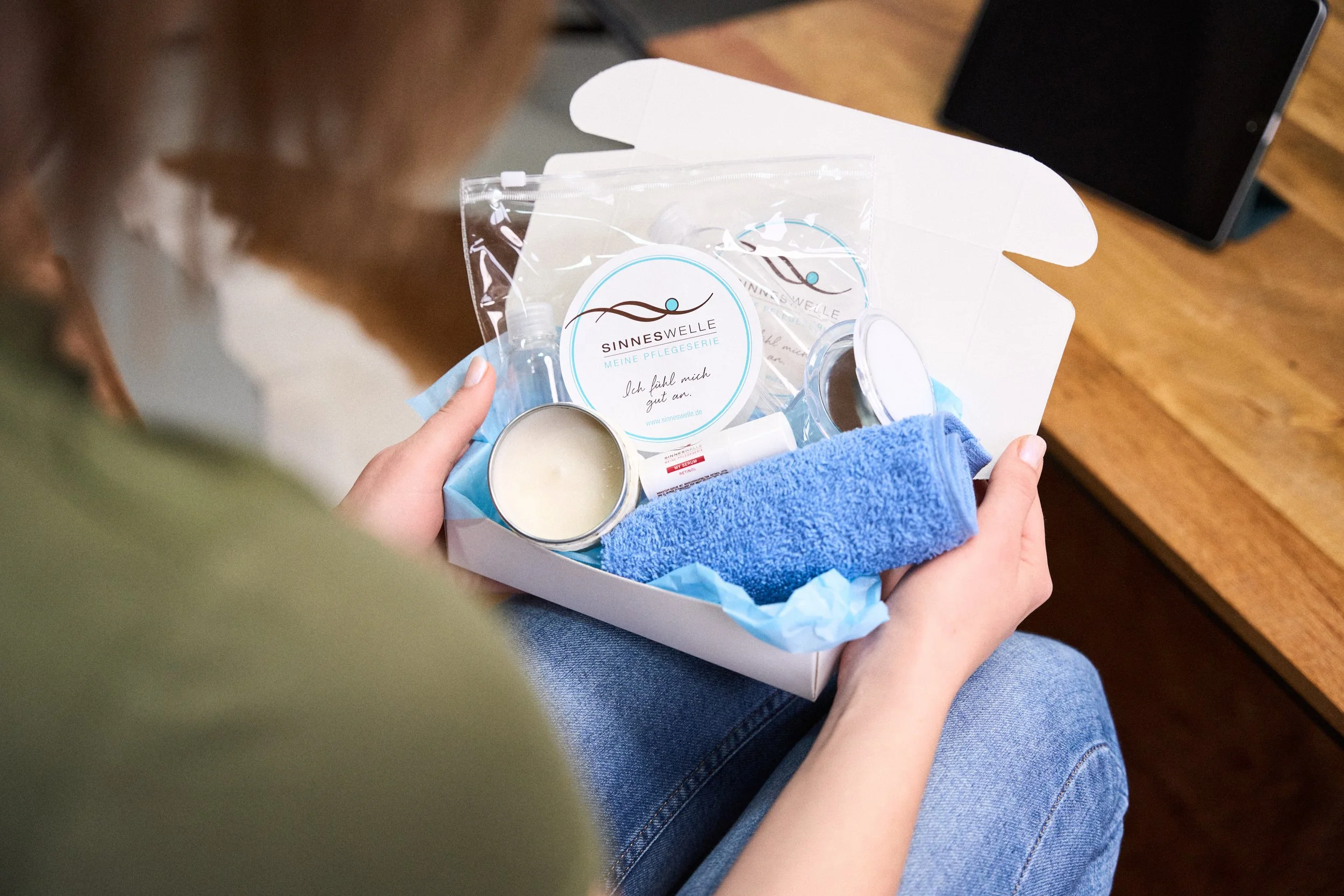 Eine Person hält ein Geschenkset mit Pflegeprodukten, einem Handtuch, einer Kerze und Pflegeartikeln in einer Schachtel, auf einem Holzstuhl sitzend.