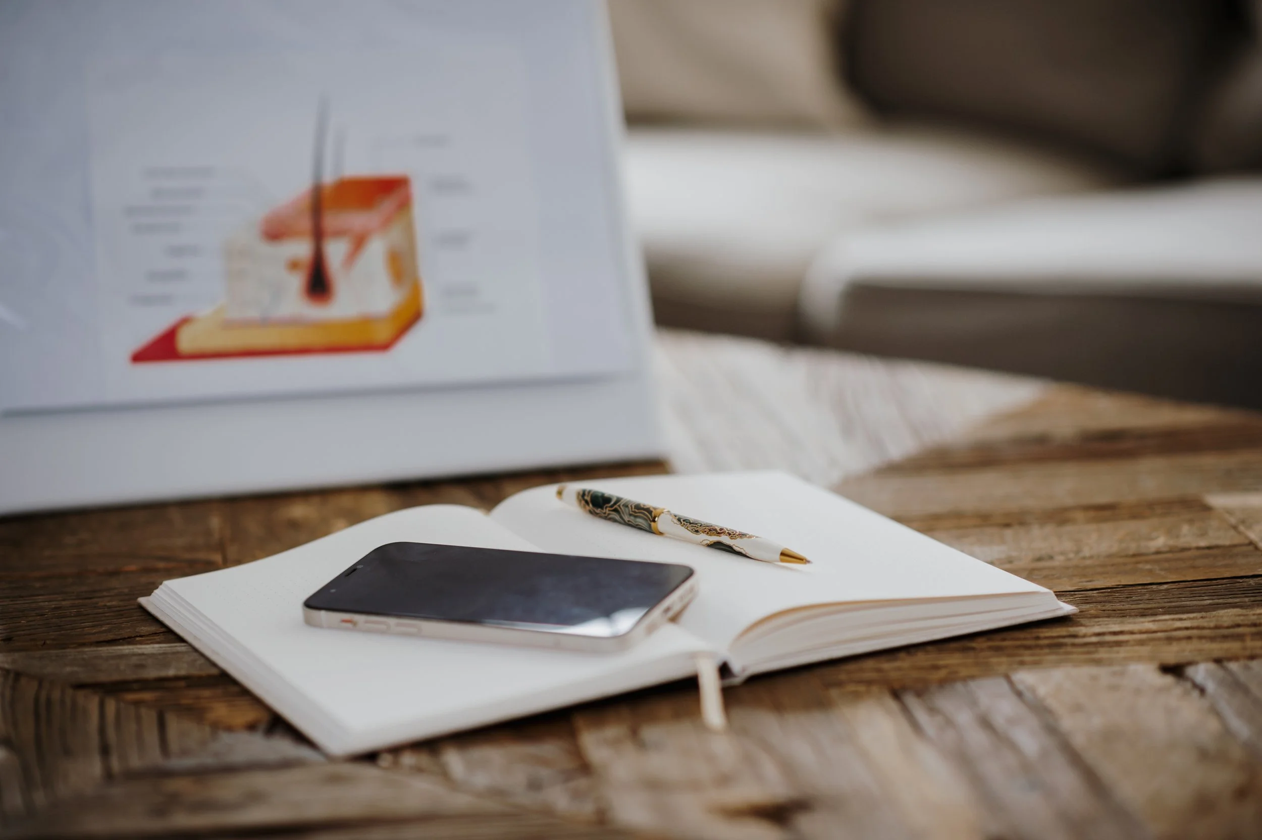 Ein aufgeschlagenes Notizbuch mit einem Stift und einem Smartphone auf einem Holzoberfläche; im Hintergrund eine Waage auf einem Papier
