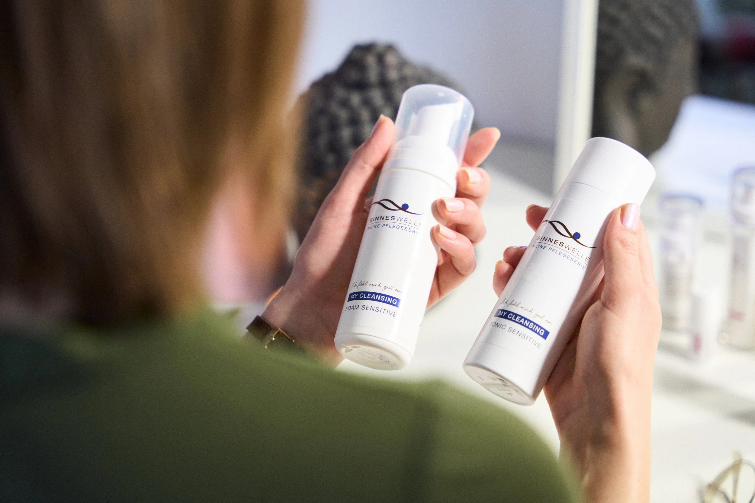 Eine Frau hält zwei weiße Hautpflegeprodukte mit blauer und schwarzer Beschriftung in den Händen, während sie sich in einem Spiegel betrachtet, im Hintergrund sind weitere Pflegeprodukte sichtbar.