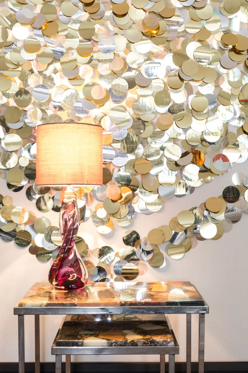 A decorative wall with metallic gold and silver circular discs, a table with a marble top, a pink and purple glass lamp, and a smaller marble-topped table underneath.