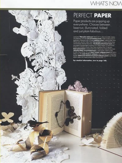 Display of white paper flowers, a book with a beaded necklace, a ribbon, and paper scraps on a table with black and white background featuring paper art.