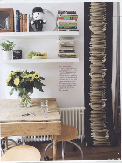A cozy corner with a wooden table, a glass of water, a flower bouquet, and a small chair. There are white shelves above with books, a small plant, and a decorative item. To the right, a tall black bookshelf is filled with stacked papers or files. A r