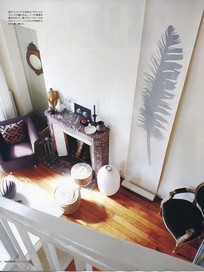 Interior of a cozy living area with a dark armchair, a marble fireplace, a tall mirror, decorative items on the mantle, an oval ottoman, a large white ceramic vase, and a shadow of a fern leaf cast on a sliding door.