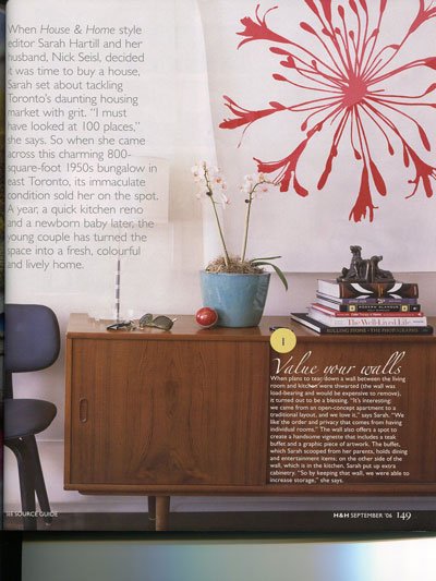 A mid-century modern wooden sideboard with black legs, topped with a blue ceramic pot containing a tall, thin plant, a pair of sunglasses, and a small red object. A black chair is partially visible to the left. Behind the sideboard, there is a large 