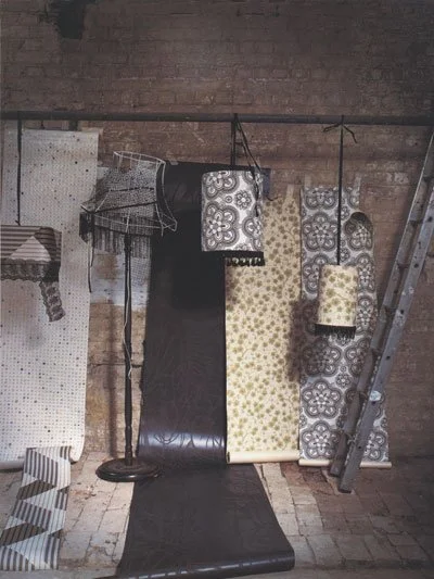 Various rolls of wallpaper with different patterns and colors hang against a brick wall, with a ladder leaning on the right side of the wall and a lamp and wire basket on the left.
