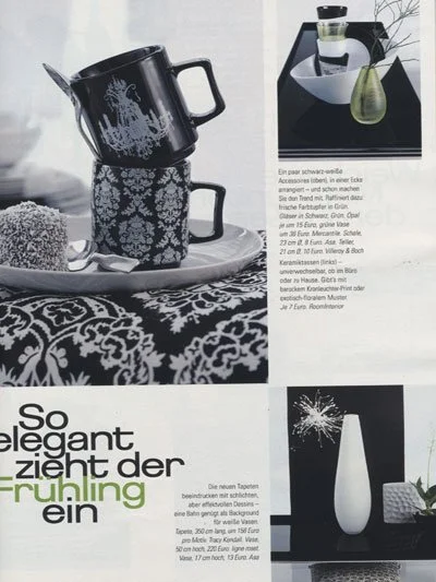 A black teapot with a floral design, a matching cup, and a silver spoon on a tray, set on a table with a black and white patterned tablecloth. There are also images of a perfume bottle, a white vase with a splash design, and some text in German.
