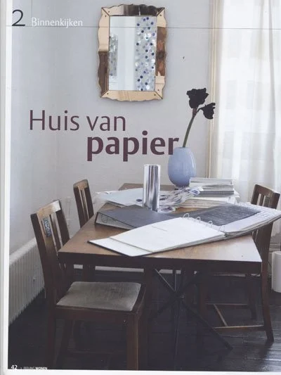 A wooden dining table with chairs, papers, and a tall tumbler in a room with a mirror, a vase with flowers, and the words 'Huis van papier' on the wall.