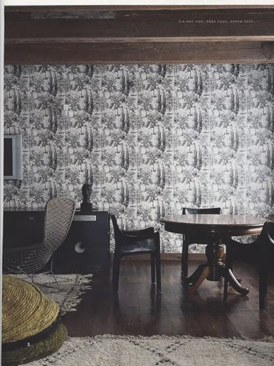 Interior of a room with a patterned wallpaper, wooden beams, a round table with four chairs, and a chair with a cushion.