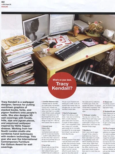 A cluttered office desk with stacks of papers and folders, a computer setup with monitor, keyboard, and mouse, and various decorative items including a skull and a helicopter model. The desk features a red circular sticker promoting Tracy Kendall.