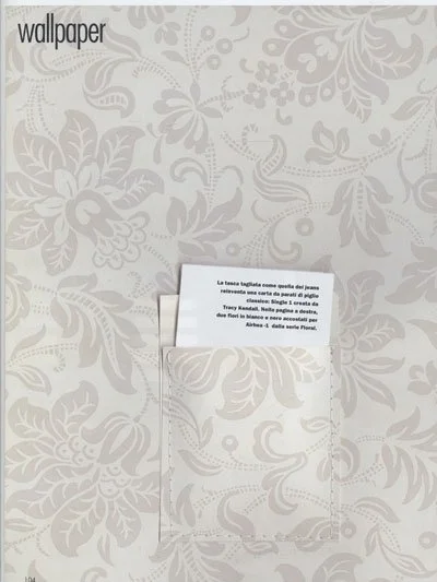 A white wall with a floral pattern, featuring a loosely held piece of paper with printed text partially visible, placed in a matching floral-patterned envelope.