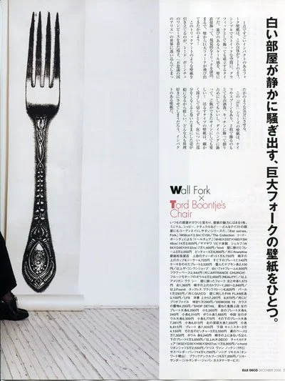 A large black and white photo of a fork with an ornate handle, leaning against a white wall next to a rolled-up piece of paper or poster on a wooden floor.