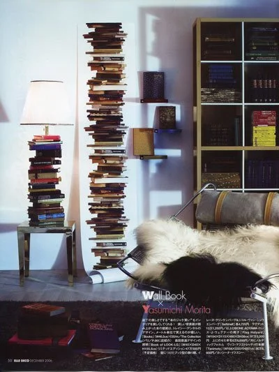 A living room with a tall, narrow bookshelf made of stacked books, a side table with more books, a tall floor lamp, a couch with furry and patterned pillows, and a square bookcase filled with books and decorative items.