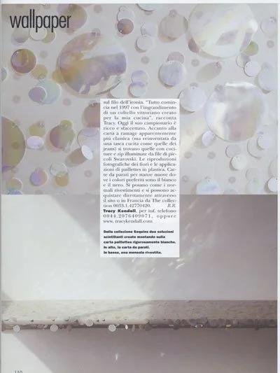 A close-up of a wall with numerous small holographic circular stickers reflecting light, creating a colorful pattern. There is a white rectangular label with text in Italian on the wall.