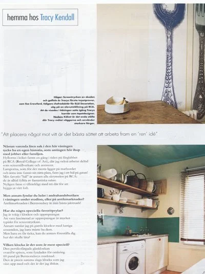 A photograph of a kitchen with a wooden countertop, washing machine, wall shelves holding dishes, a window with blinds, and decorative wall art.