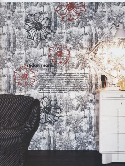 Interior of a room featuring a textured wall with black and red floral stickers, a black upholstered chair, and a white dresser with a wall-mounted lamp.
