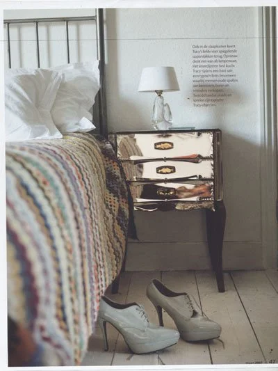 A bedroom scene with a bed that has a colorful crocheted blanket and white pillows, a mirrored nightstand with a lamp and a rifle, a pair of white high-heeled shoes on the floor, and a section of a wall with some text on it.