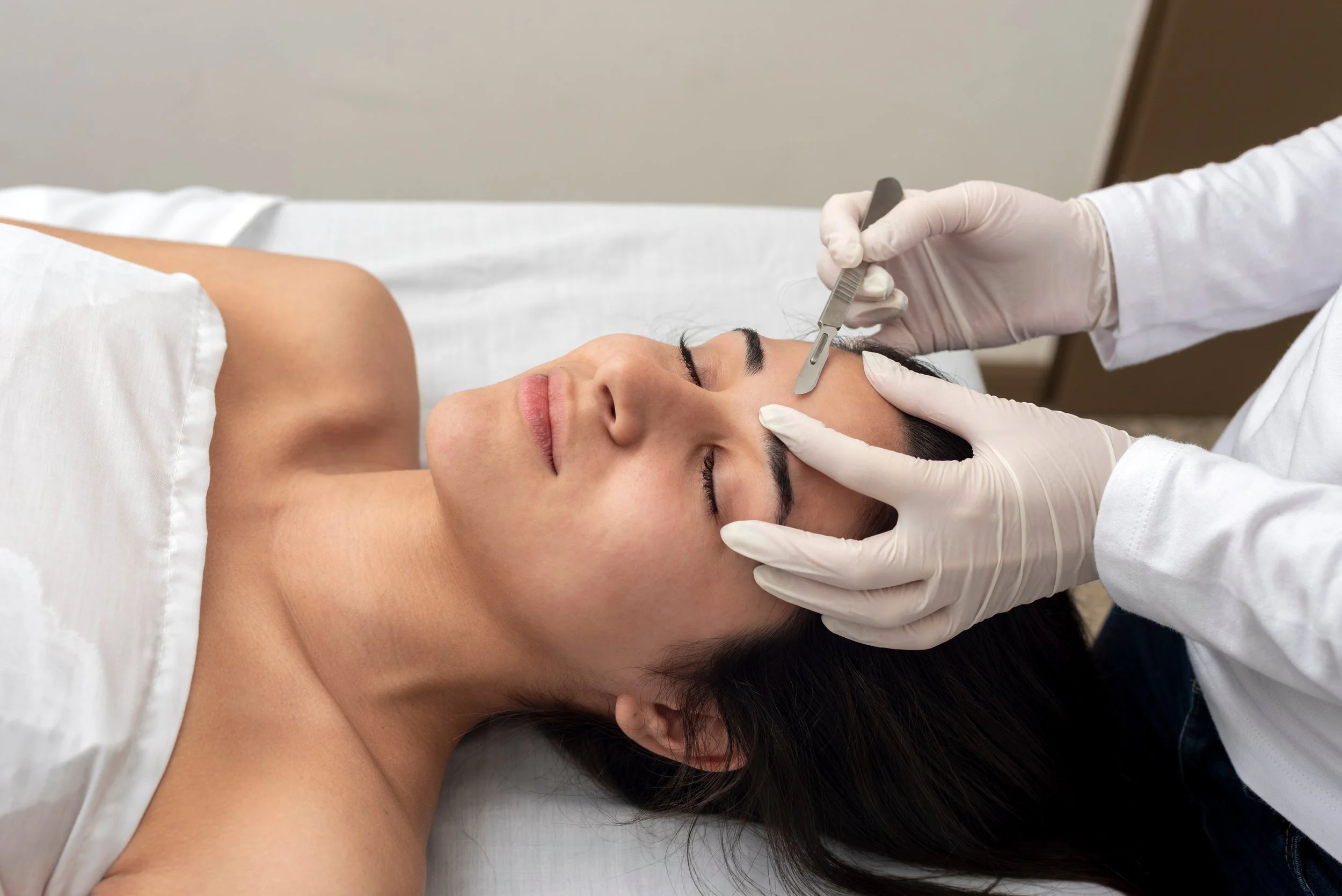 A woman lying down with her eyes closed while a doctor performs an eyebrow microblading procedure using a small handheld tool.