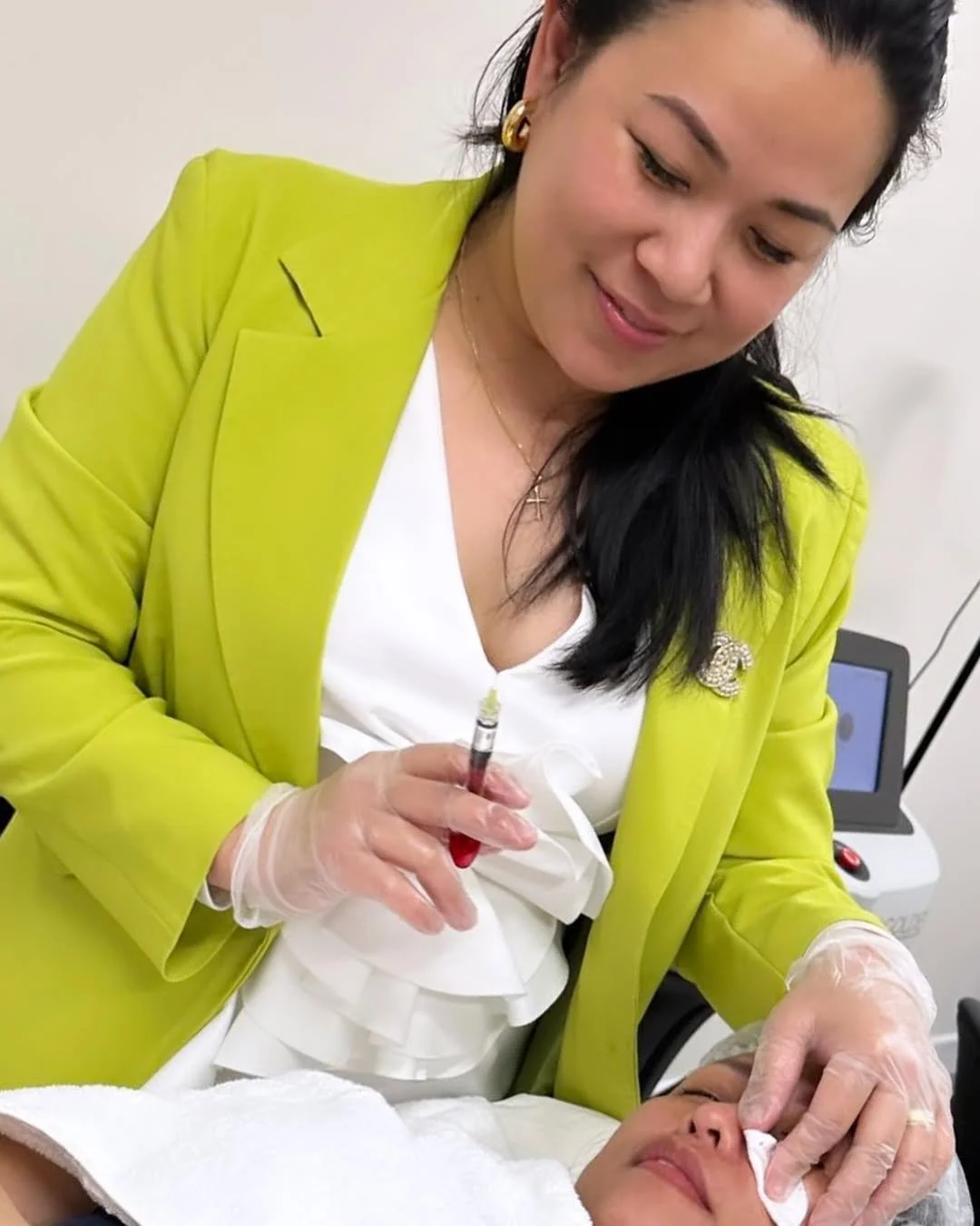 A woman in a bright yellow blazer and white dress is administering a cosmetic injection to a woman lying down, with a syringe in her right hand and a cotton swab in her left hand.