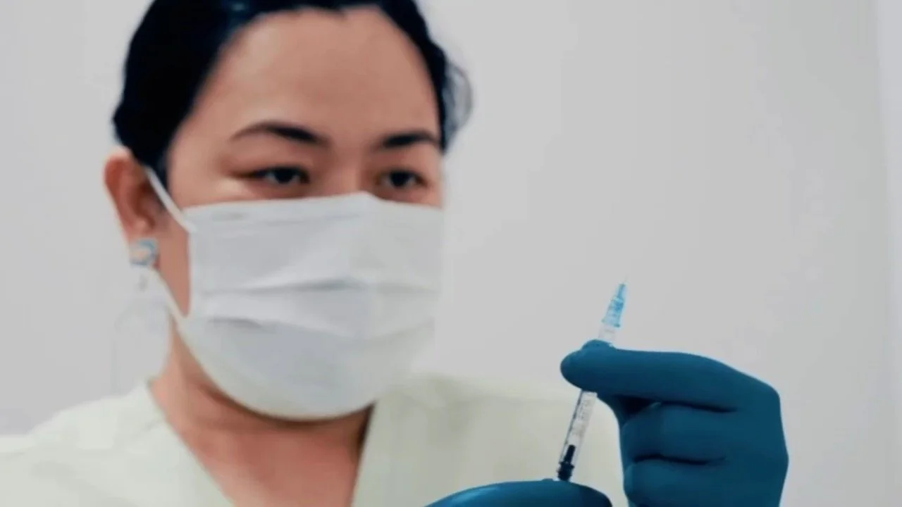 A healthcare worker wearing a face mask and gloves prepares a syringe with a vaccine or medication.
