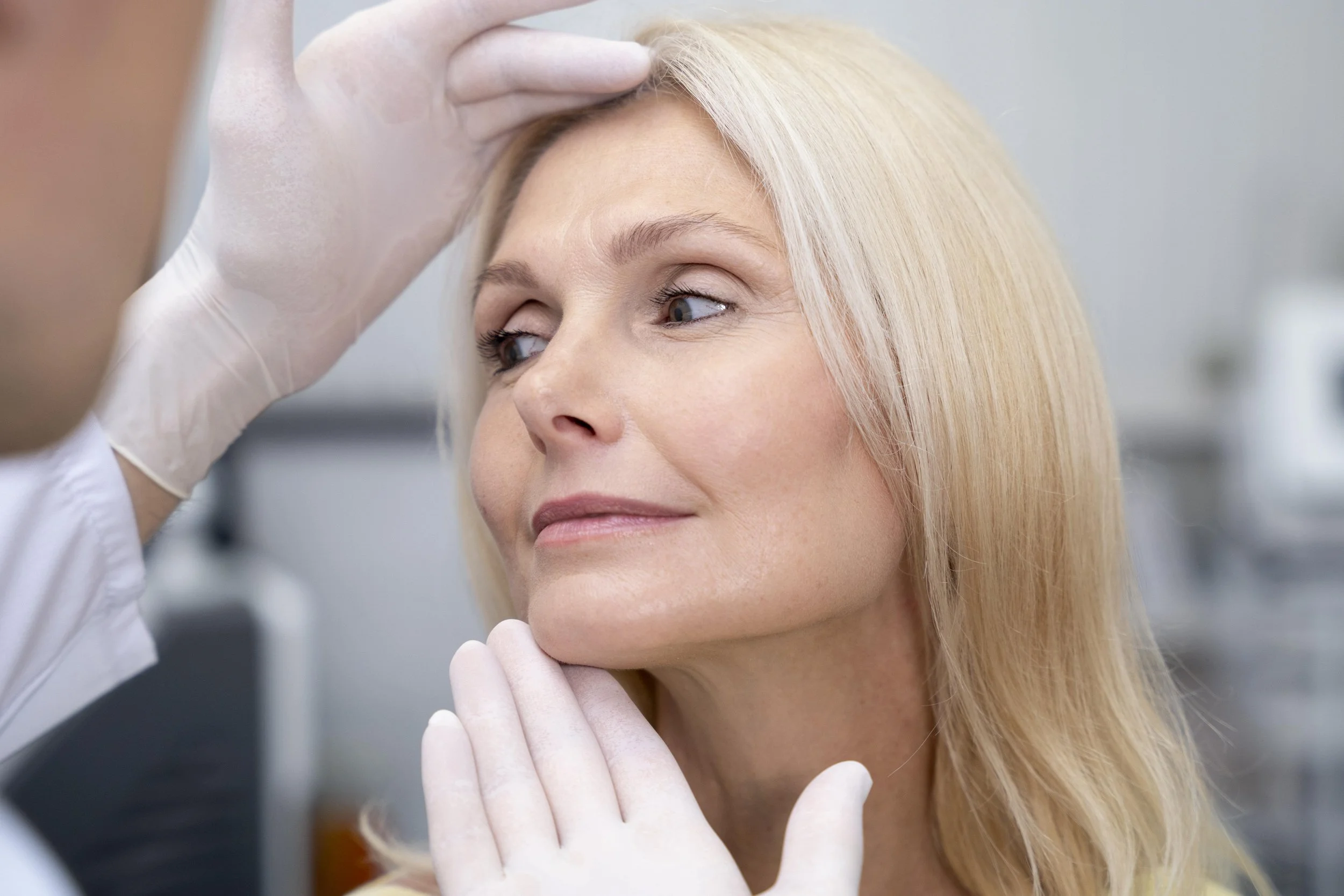 A woman having a skin check by a healthcare professional, with the professional examining her forehead and the woman holding her chin.