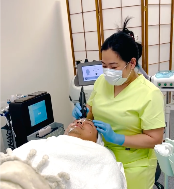 A healthcare professional in yellow scrubs, wearing a face mask and gloves, performs a cosmetic procedure on a patient's face in a clinic or spa setting.