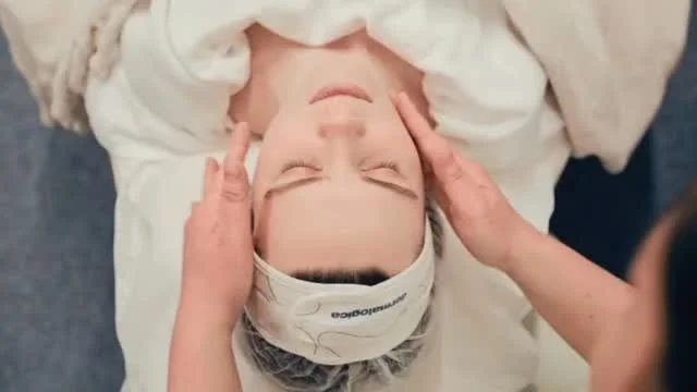 Person receiving a facial massage while lying down with eyes closed, wearing a headband.